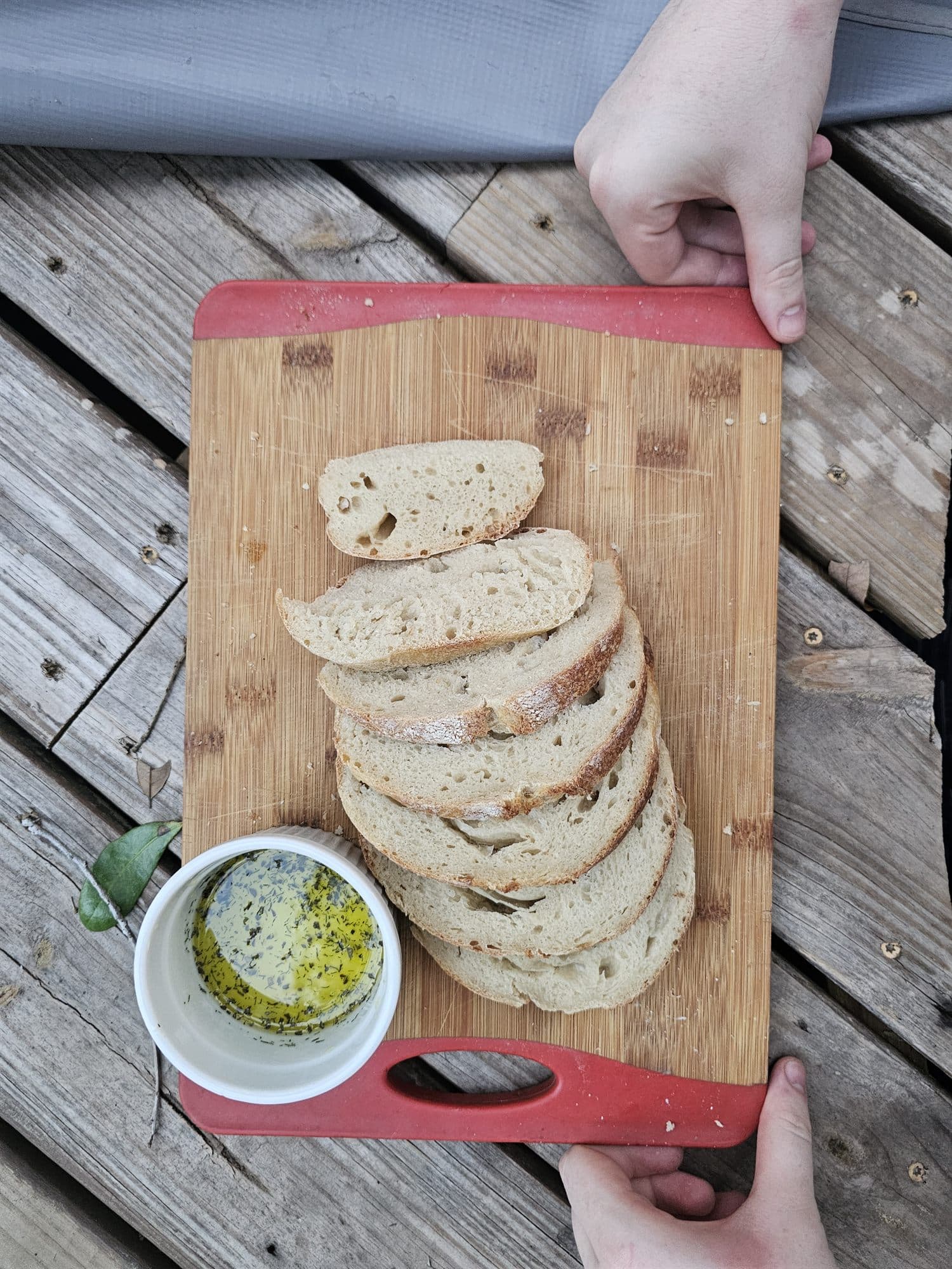 Two hands setting down a board of sliced sourdough and herbed olive oil on weathered deck boards